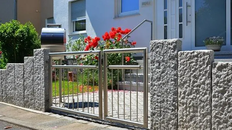 Eingangsbereich eines modernen Hauses mit Steinfassade, Edelstahlzaun und roten Blumen.