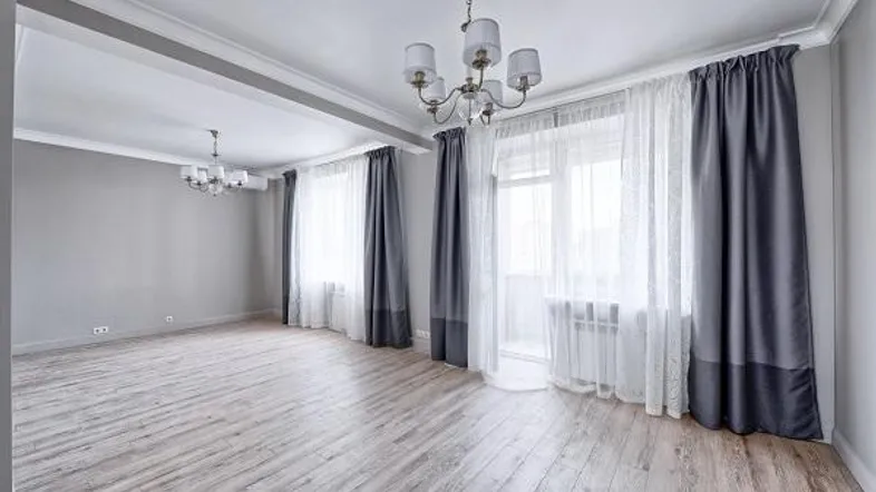 Helles, leeres Wohnzimmer mit hellem Laminatboden, großen Fenstern und eleganten Vorhängen.