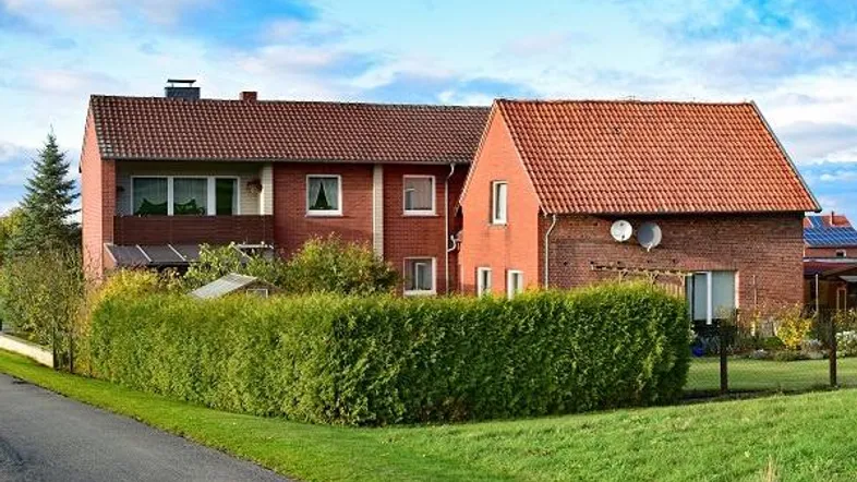 Zwei Einfamilienhäuser mit roten Ziegeldächern und gepflegter Hecke, umgeben von Grünflächen. Musterfoto.