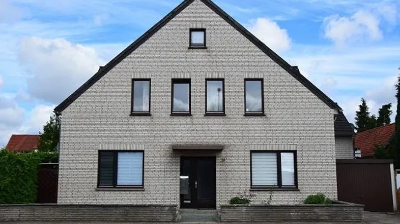 Einfamilienhaus mit heller Ziegelfassade, Satteldach und kleinen Fenstern unter bewölktem Himmel.