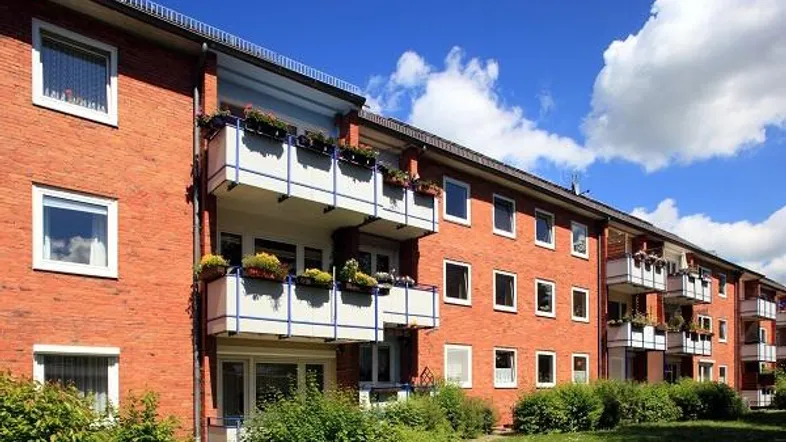 Mehrfamilienhaus mit roter Backsteinfassade, Balkonen und grüner Umgebung unter blauem Himmel.