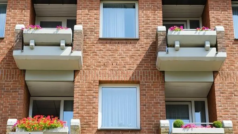 Ziegelsteinfassade eines Mehrfamilienhauses mit mehreren Balkonen, die mit bunten Blumen geschmückt sind.