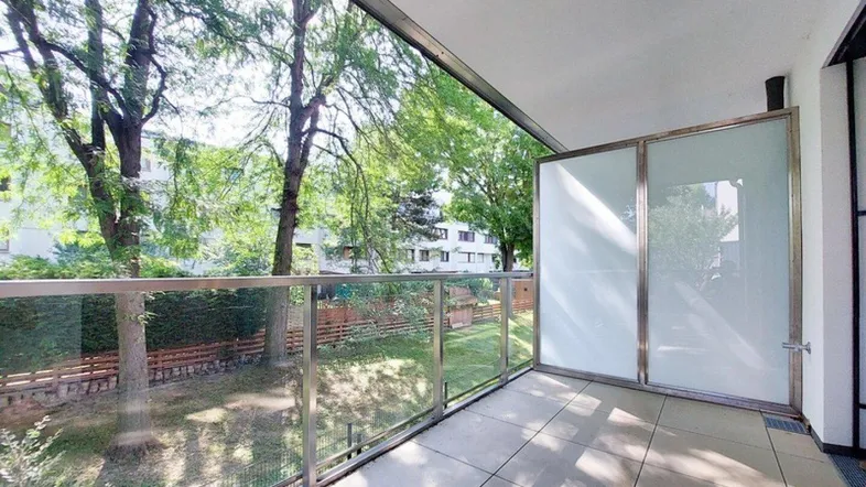 Heller Balkon mit Glasgeländer, Fliesenboden und Blick ins Grüne mit Bäumen.
