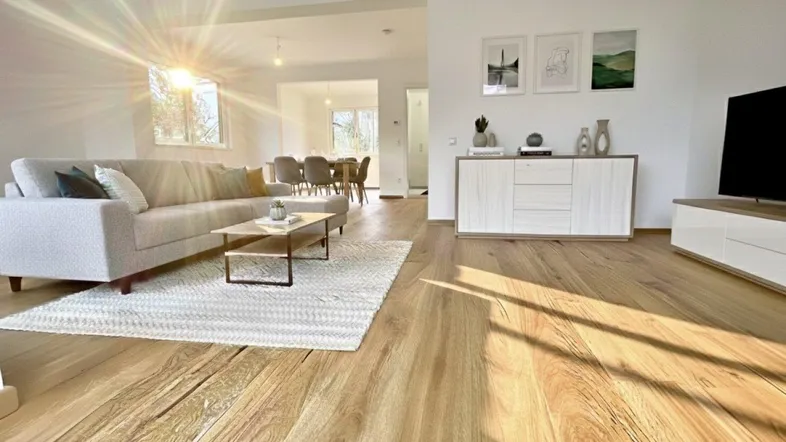 Helles, offenes Wohnzimmer mit Holzboden, modernem Sofa und Essbereich im Hintergrund, durchflutet von Sonnenlicht.