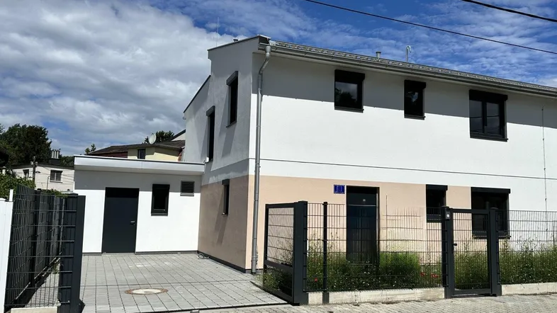 Moderne Hausfassade mit weißem und beigem Putz, dunklen Fenstern und gepflasterter Einfahrt.