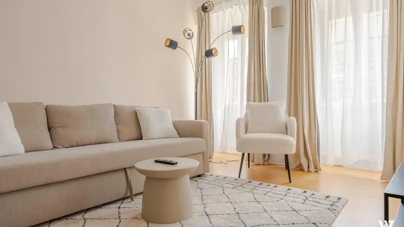 Helles Wohnzimmer mit beigem Sofa, modernem Teppich und großen Fenstern mit Vorhängen.