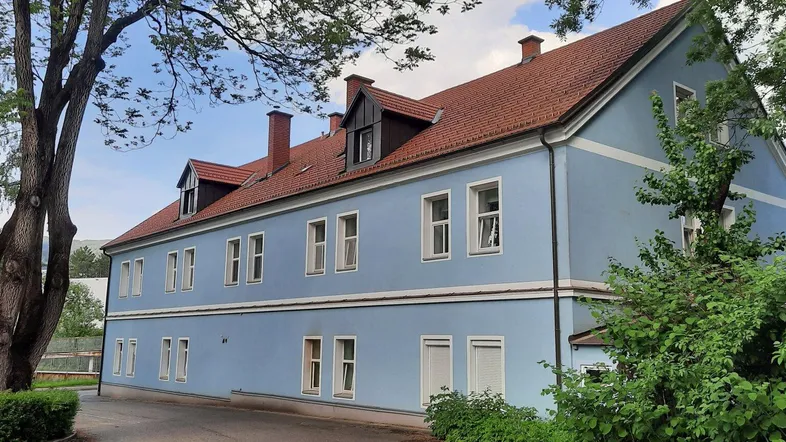 Ansicht des hellblauen Gebäudes mit rotem Ziegeldach und umgebenden Bäumen unter blauem Himmel.