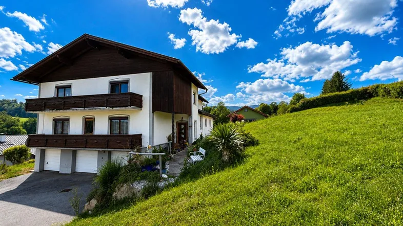 Traditionelles Haus mit weißer Fassade und Holzbalkonen, eingebettet in eine grüne Hügellandschaft unter blauem Himmel.