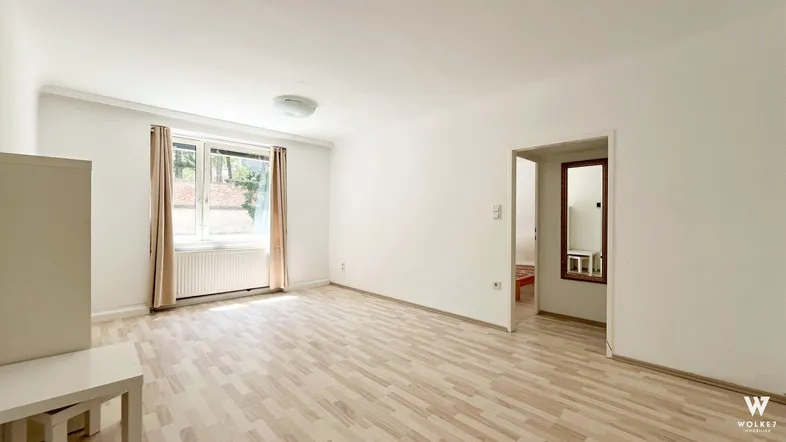 Heller, leerer Wohnraum mit Laminatboden und großem Fenster, das viel Tageslicht hereinlässt.