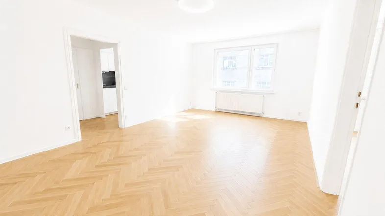 Helles, leeres Wohnzimmer mit Fischgrätparkettboden und weißen Wänden, bereit zur Einrichtung.