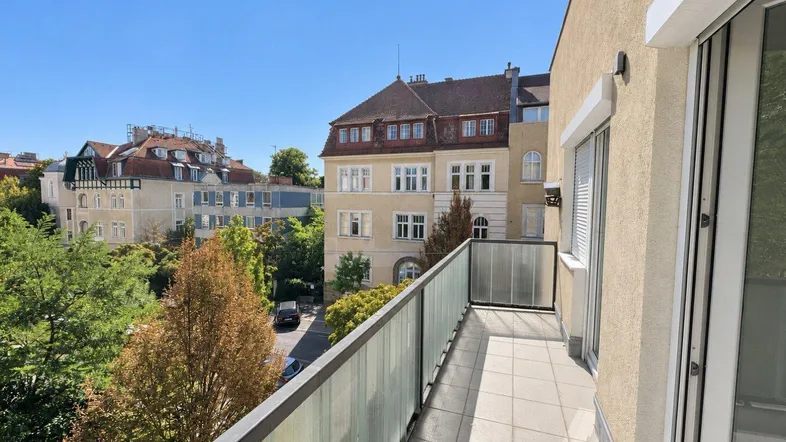 Heller Balkon mit Fliesenboden und Glasgeländer, bietet einen Blick auf die umliegenden Gebäude und Bäume.