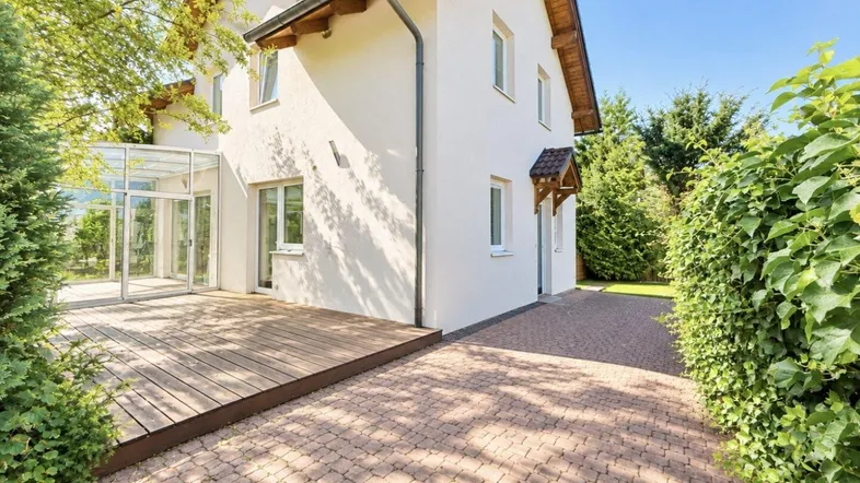Modernes weißes Einfamilienhaus mit Holzterrasse, gepflasterter Auffahrt und Wintergarten.