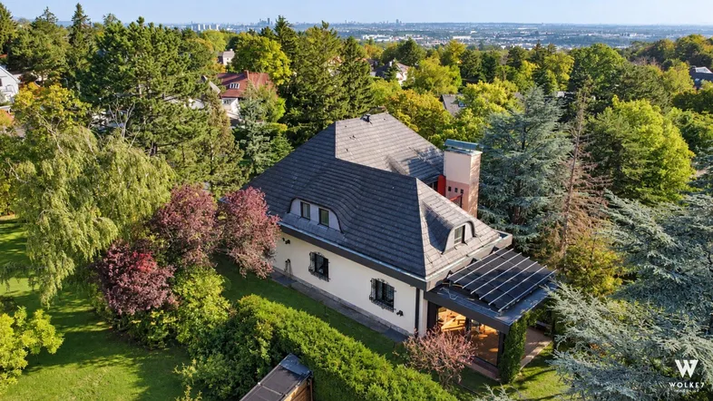 Luftaufnahme eines Hauses mit dunklem Ziegeldach, umgeben von üppigem Grün und Bäumen, mit Stadtblick in der Ferne.