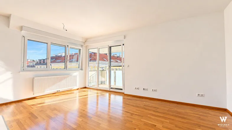 Helles Zimmer mit Parkettboden, weißen Wänden und Zugang zu einem sonnigen Balkon.