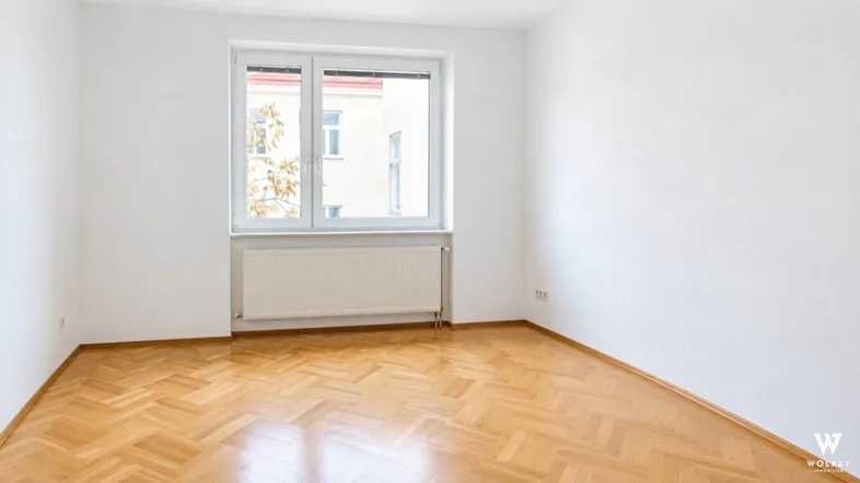 Heller Raum mit Fischgrätparkettboden, weißen Wänden und einem Fenster mit Blick auf ein Gebäude.