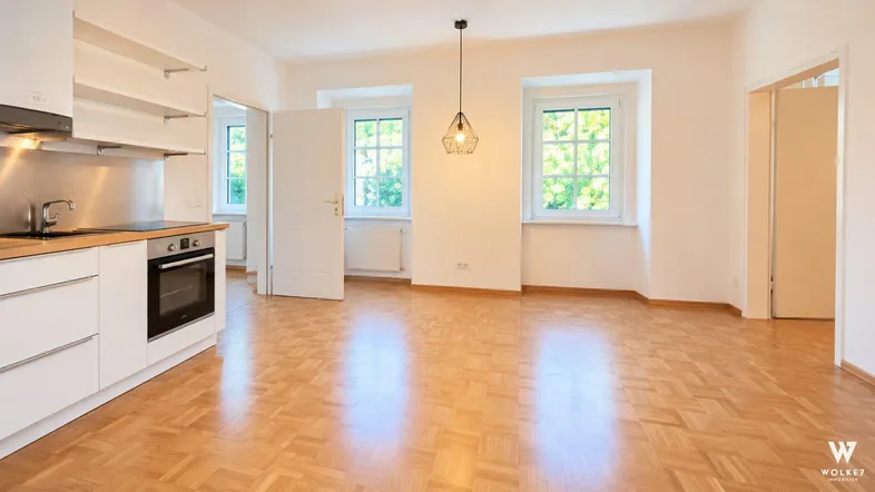 Helles Wohnzimmer mit integrierter Küchenzeile, Parkettboden und zwei großen Fenstern.