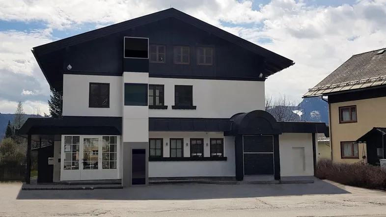 Traditionelles Gebäude mit dunklem Dach und weißer Fassade in alpiner Umgebung.
