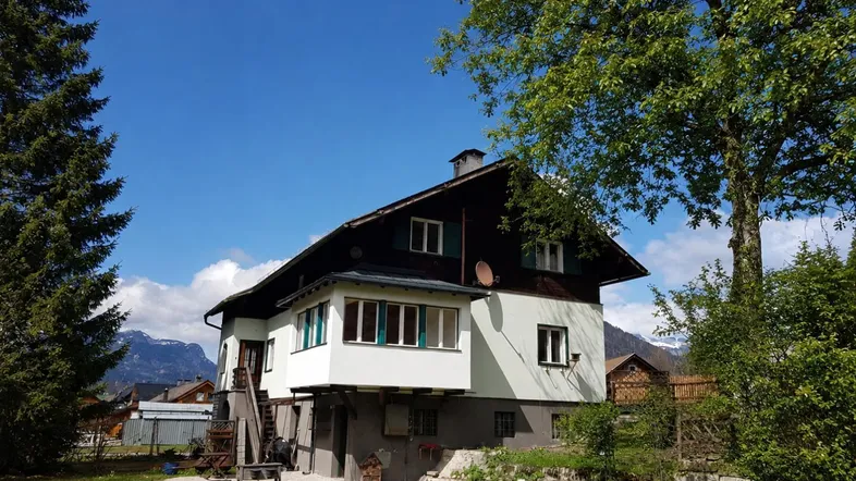 Traditionelles Haus mit weißer Fassade, Holzbalkonen und einem grünen Gartenbereich unter blauem Himmel.