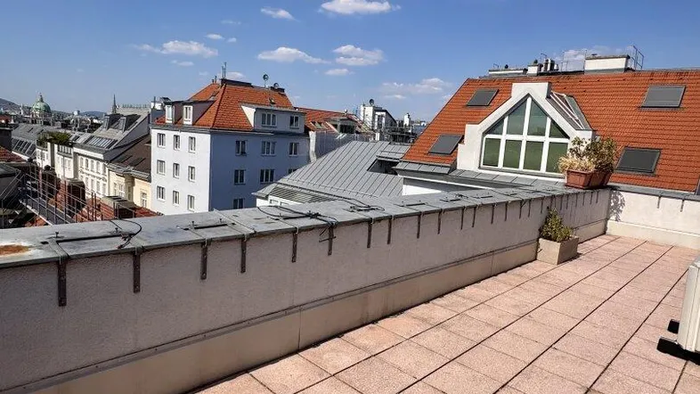 Geräumige Dachterrasse mit Fliesenboden und weitem Blick über die Dächer der Stadt unter blauem Himmel.