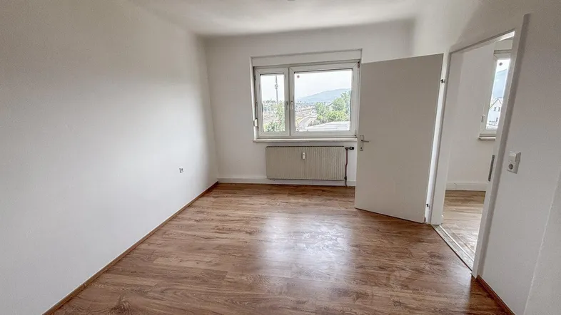 Helles, leeres Zimmer mit Laminatboden und Fensterblick ins Grüne.