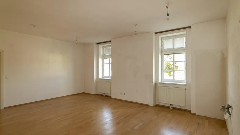 Helles, leeres Zimmer mit Holzfußboden und zwei Fenstern, bereit zur individuellen Gestaltung.