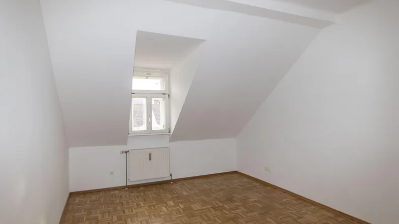 Helles Zimmer im Dachgeschoss mit Dachschräge, Fenster und Parkettboden.