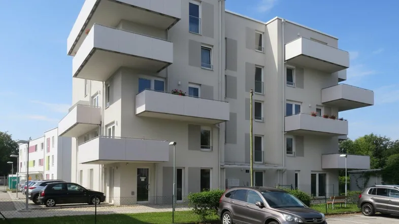 Modernes Mehrfamilienhaus mit hellgrauer Fassade, Balkonen und Außenparkplätzen unter blauem Himmel.