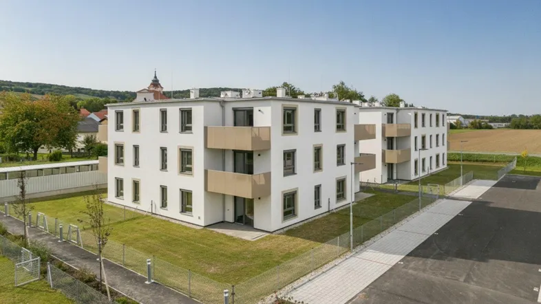 Moderne Neubauten mit weißer Fassade und Balkonen, umgeben von Grünflächen und Parkplätzen unter blauem Himmel.