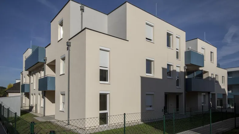 Modernes Mehrfamilienhaus mit heller Fassade und blauen Balkonen unter blauem Himmel.