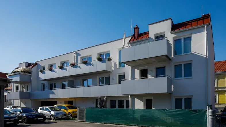 Modernes Mehrfamilienhaus mit Balkonen und Parkplätzen unter blauem Himmel.