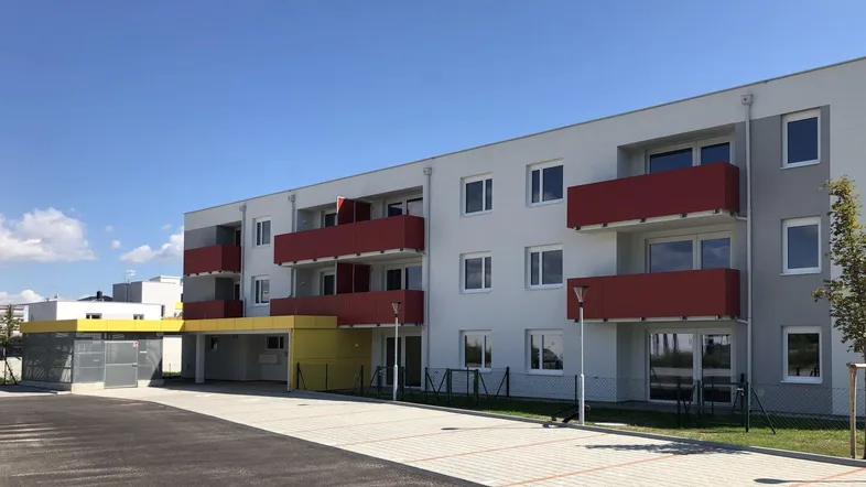 Modernes Mehrfamilienhaus mit roten Balkonen und Parkplätzen unter blauem Himmel.