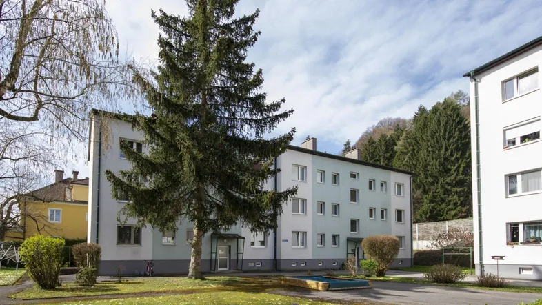 Mehrfamilienhaus mit hellgrauer und hellgrüner Fassade, umgeben von Grünflächen und Bäumen unter blauem Himmel.