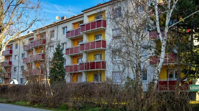 Mehrfamilienhaus mit farbigen Balkonen und umgebender Grünfläche unter blauem Himmel.