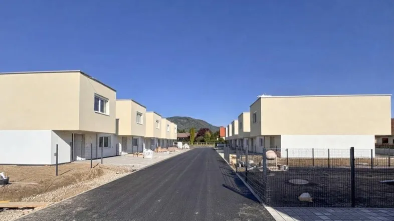 Neue Reihenhäuser in einer Siedlung mit asphaltierter Straße und Bergblick unter blauem Himmel.