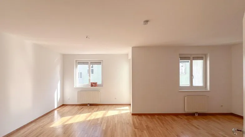 Helles, leeres Zimmer mit Parkettboden und zwei Fenstern, bereit zur individuellen Gestaltung.
