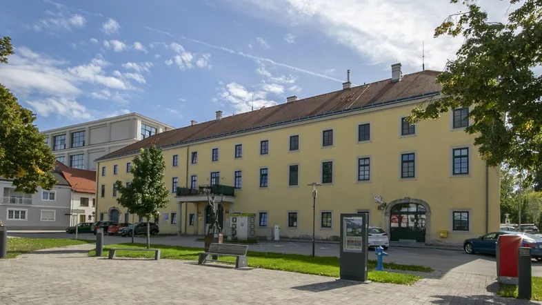 Historisches Gebäude mit gelber Fassade, blauen Fenstern und Parkplätzen im Freien unter blauem Himmel.