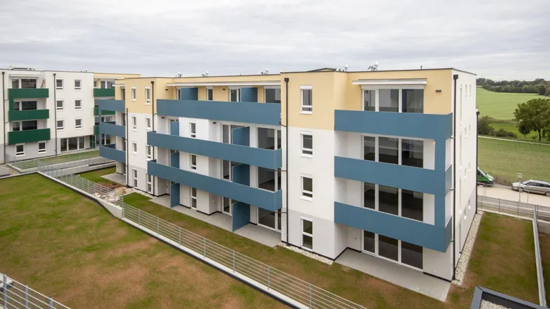 Moderne Neubauwohnanlage mit blauen Balkonen und grünen Außenbereichen unter bewölktem Himmel.