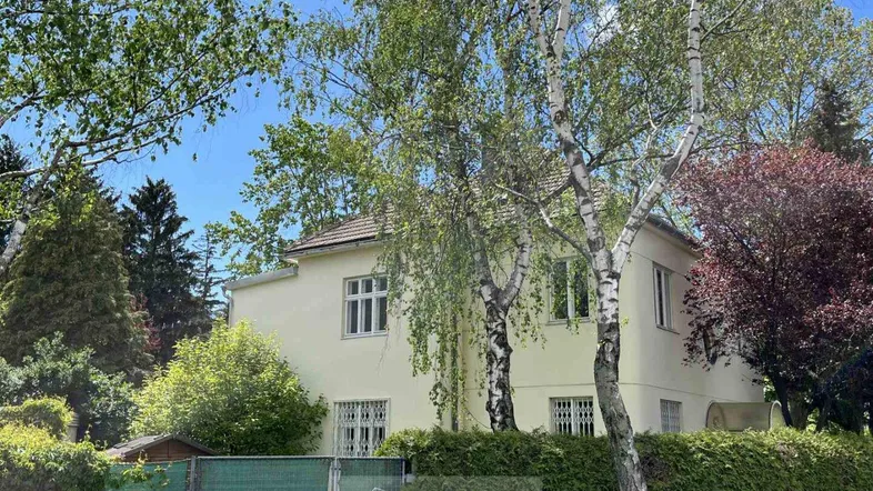 Gelbe Fassade eines Hauses umgeben von üppigem Grün und hohen Bäumen unter blauem Himmel.