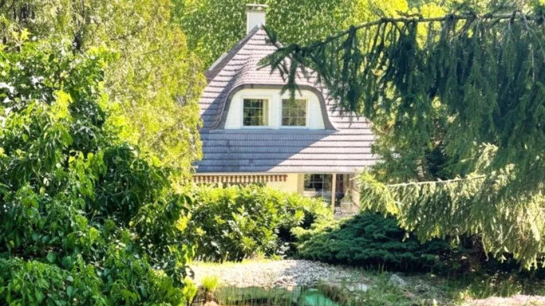 Charmantes Haus mit Mansarddach, umgeben von üppigem Grün und einem kleinen Teich.