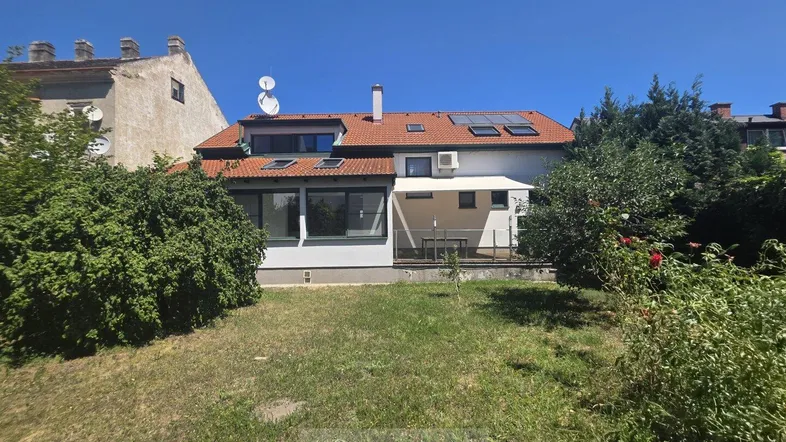 Hinteransicht des Hauses mit Garten, Wintergarten und Terrasse unter blauem Himmel.
