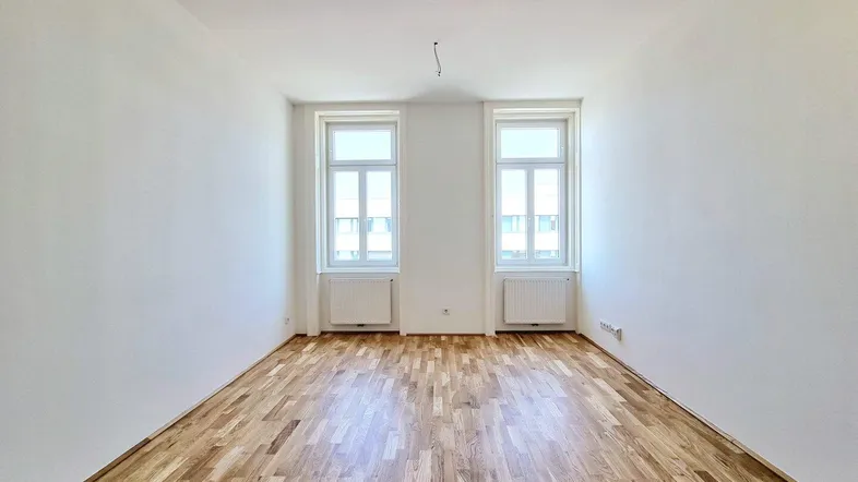 Helles, leeres Zimmer mit Parkettboden und zwei Fenstern, bereit zur individuellen Gestaltung.