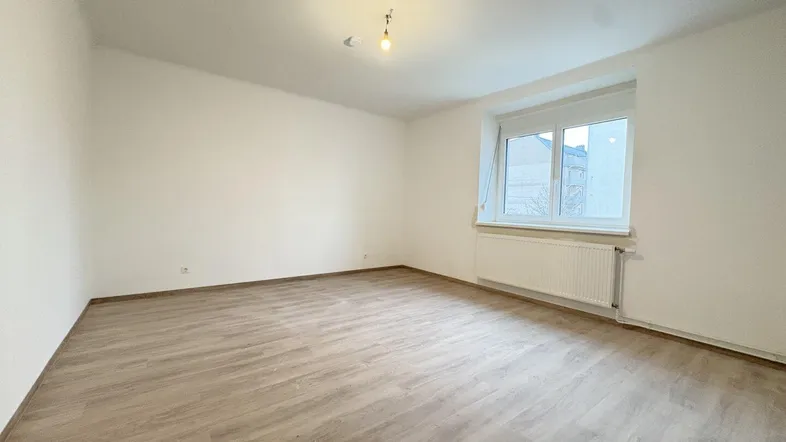 Helles, leeres Zimmer mit Laminatboden und einem Fenster, das Tageslicht hereinlässt.