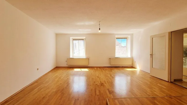 Helles, leeres Wohnzimmer mit Parkettboden und zwei Fenstern, die viel Tageslicht hereinlassen.
