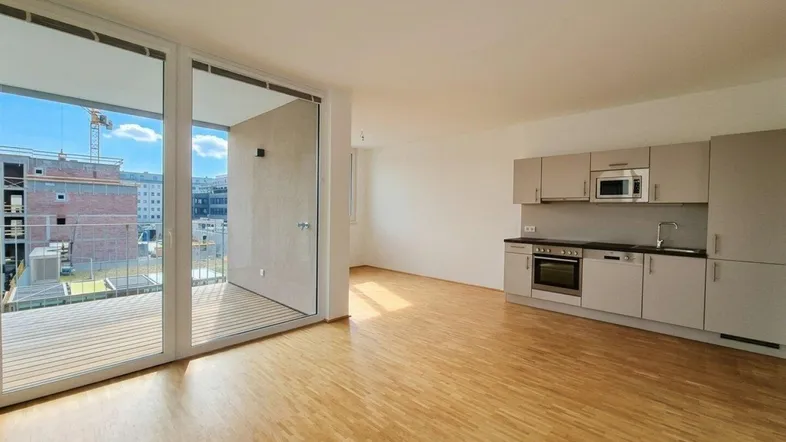 Heller offener Wohnbereich mit integrierter Küche, Parkettboden und Zugang zum Balkon.