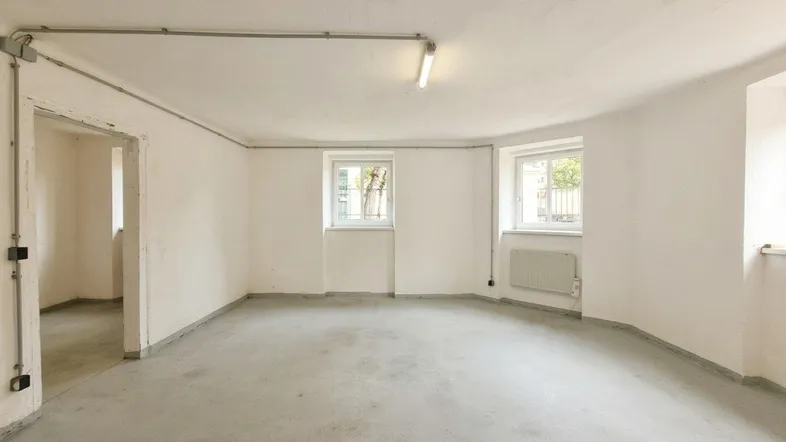 Heller Lagerraum mit zwei Fenstern, Betonboden und sichtbaren Leitungen an den Wänden.