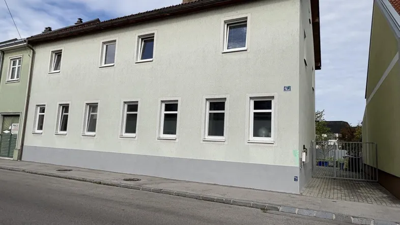 Helles Mehrfamilienhaus mit schlichter Fassade und mehreren Fenstern an einer ruhigen Straße gelegen.