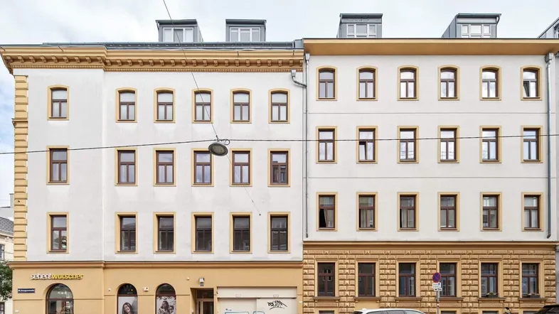 Fassade zweier Gebäude mit Geschäften im Erdgeschoss und parkenden Autos auf der Straße.