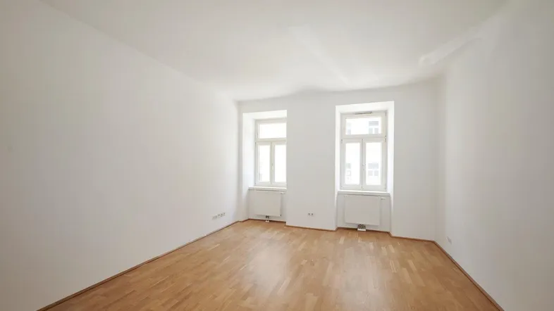 Helles, leeres Zimmer mit Parkettboden und zwei Fenstern, bereit zur individuellen Gestaltung.