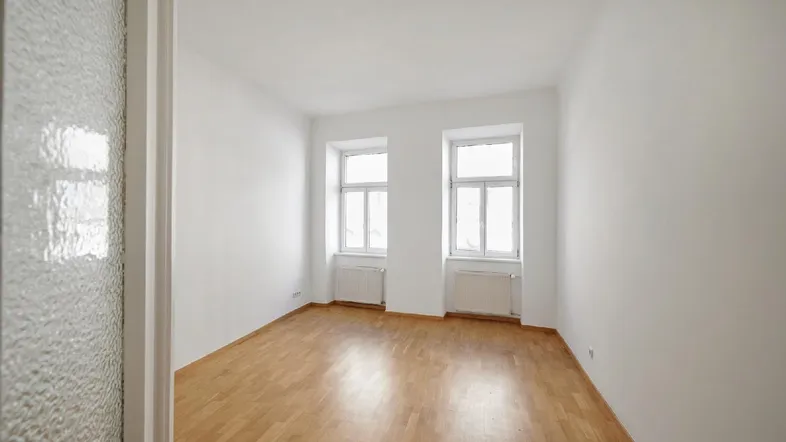 Helles, leeres Zimmer mit Parkettboden und zwei Fenstern, bereit zur individuellen Gestaltung.