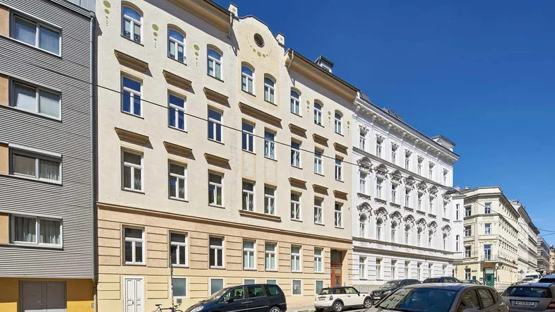 Eine historische Wohnfassade mit vielen Fenstern und parkenden Autos auf der Straße unter blauem Himmel.